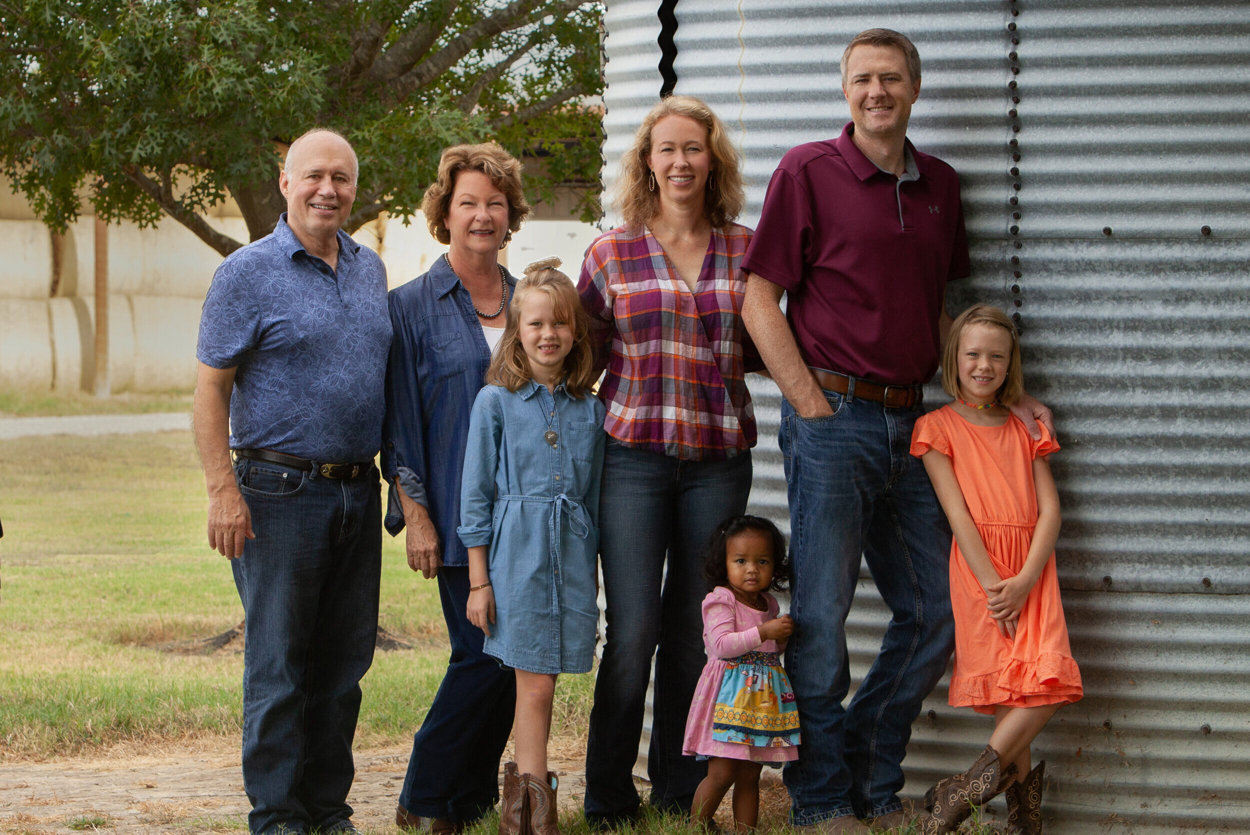 A multigenerational family portrait featuring six members standing together outdoors near a corrugated metal structure. The group includes three adults and three children, all dressed in casual attire, showcasing a warm and friendly atmosphere.