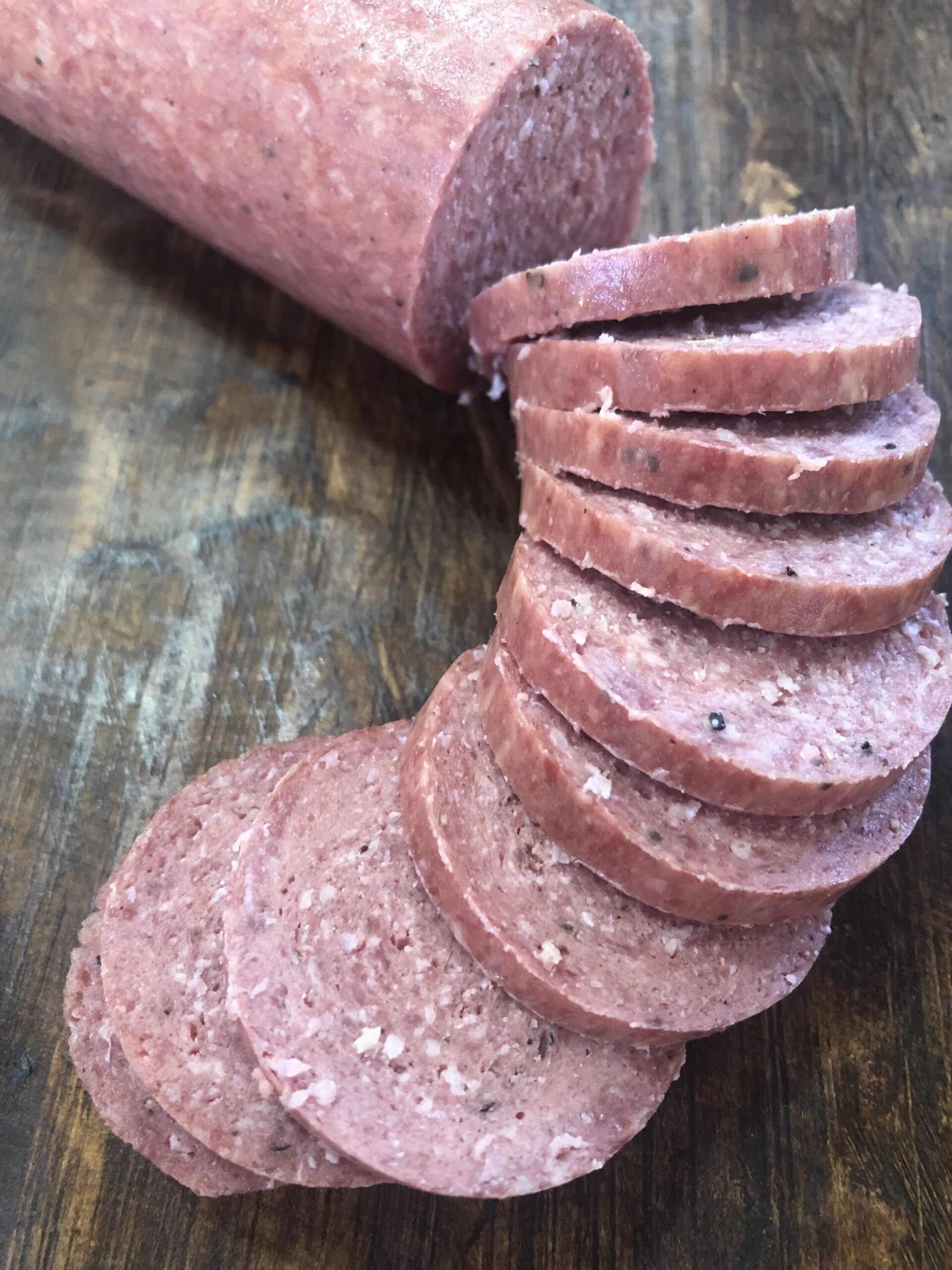 Sliced deli meat arranged on a wooden surface, showcasing a cylindrical log of processed meat with visible spices and textures, ideal for sandwiches or charcuterie boards.